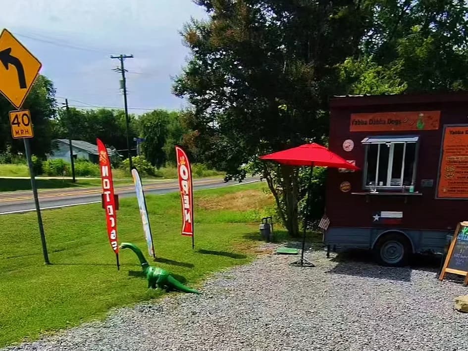 The Yabba Dabba Dogs hot dog food truck along the roadside in Nevada, Texas, marked by 'Open' and 'Hot Dogs' feather flags and a green dinosaur statue
